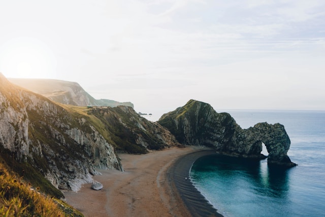 Image of a curved beach with rugged hills behind the beach and an arched tunnel through the rocks over the water